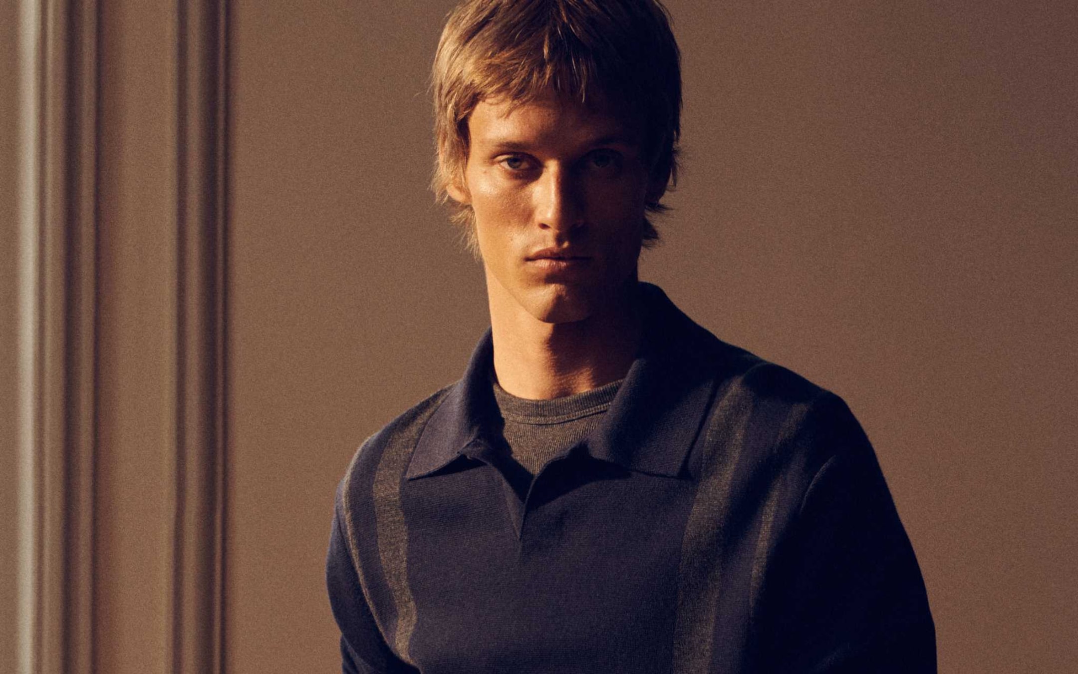 A young person with short blond hair wearing a dark collared sweater stands in soft, warm lighting against a blurred, plain background.