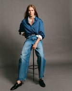 Placeholder for: Person sitting on a stool wearing a blue striped shirt, blue jeans, and black shoes, with a neutral expression and long hair.