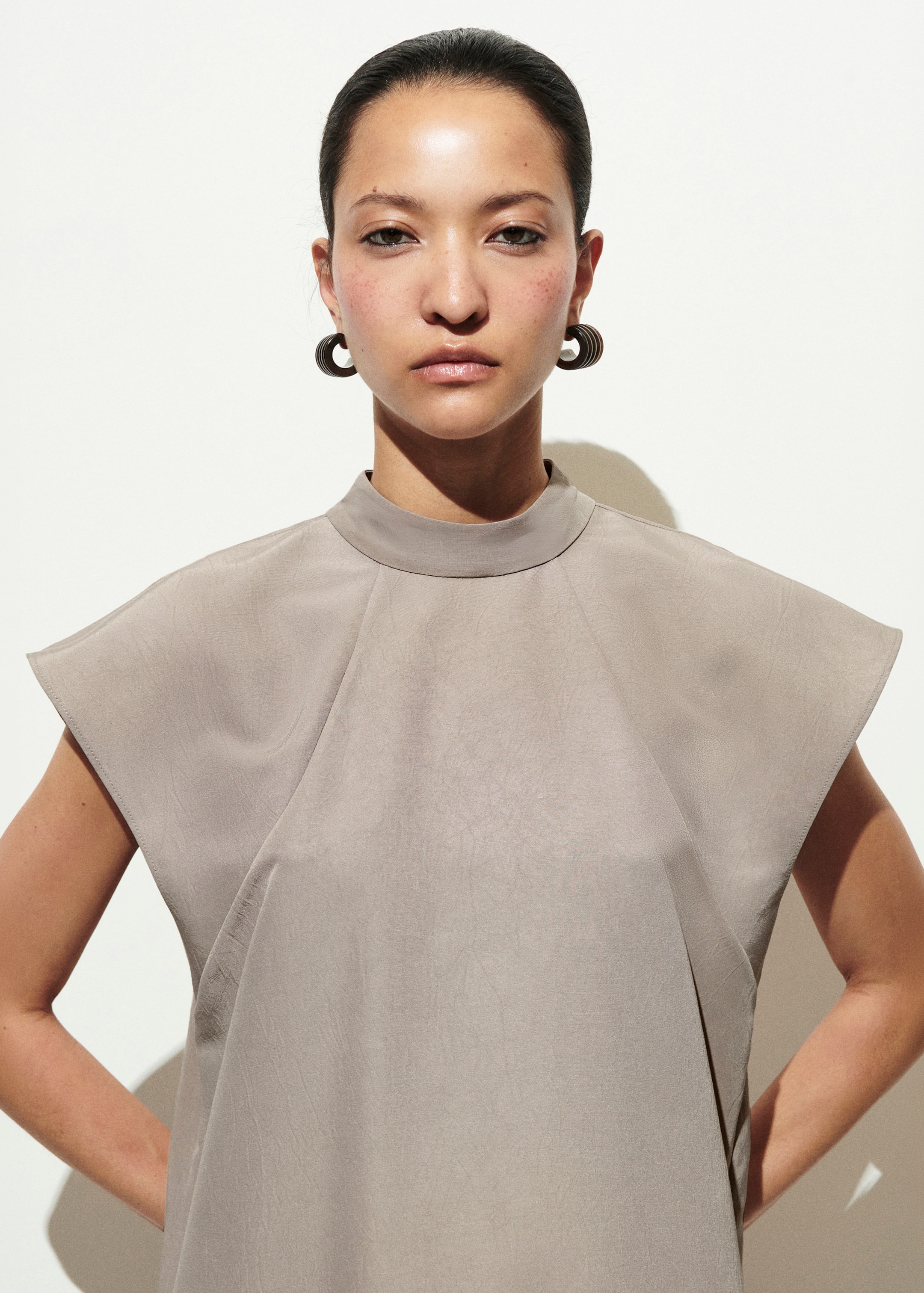 Person in a minimalist gray outfit with large hoop earrings, standing against a plain white background, hands on hips.