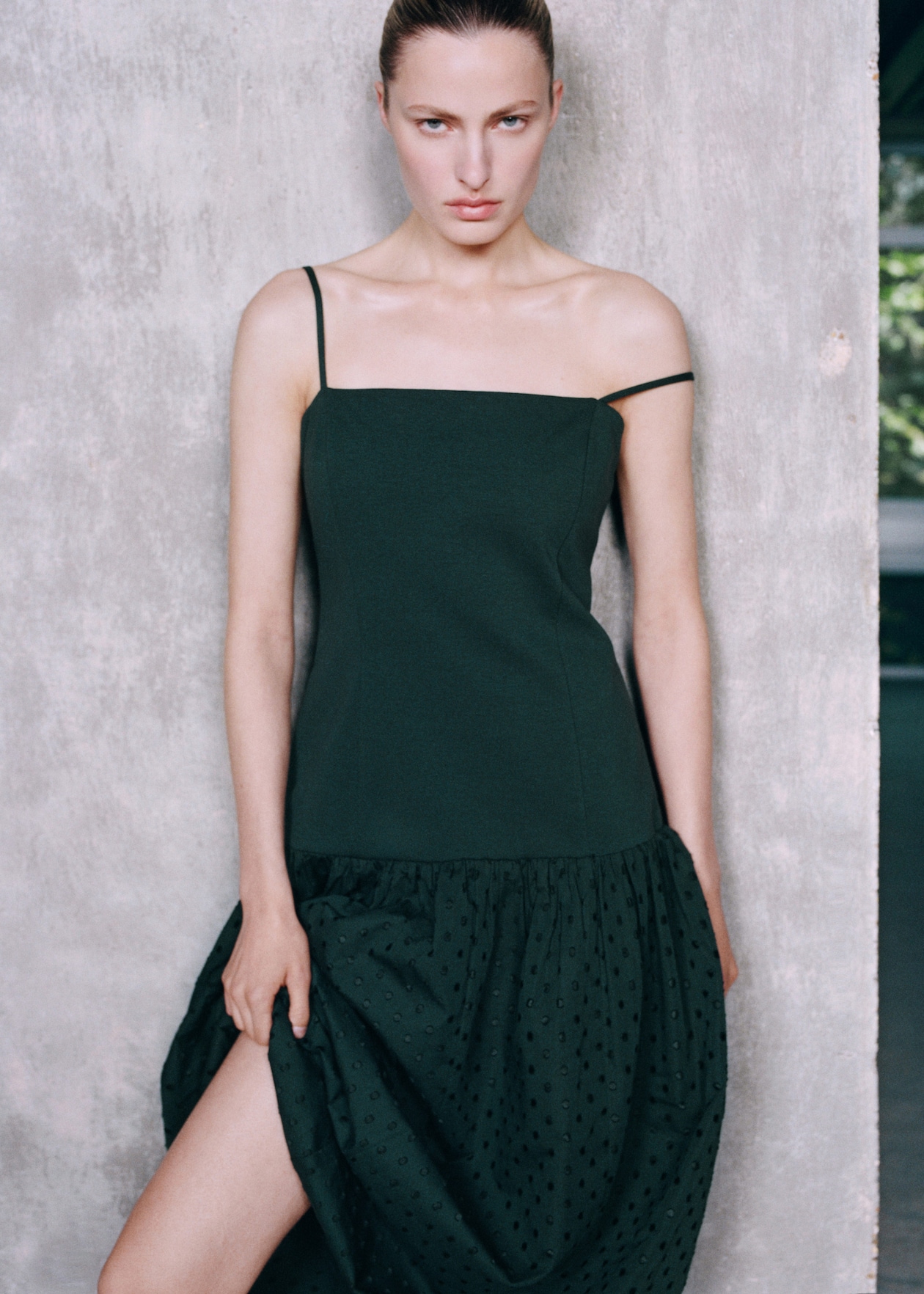 Woman in a dark green dress with thin straps, standing against a textured gray wall, holding the hem of her skirt.
