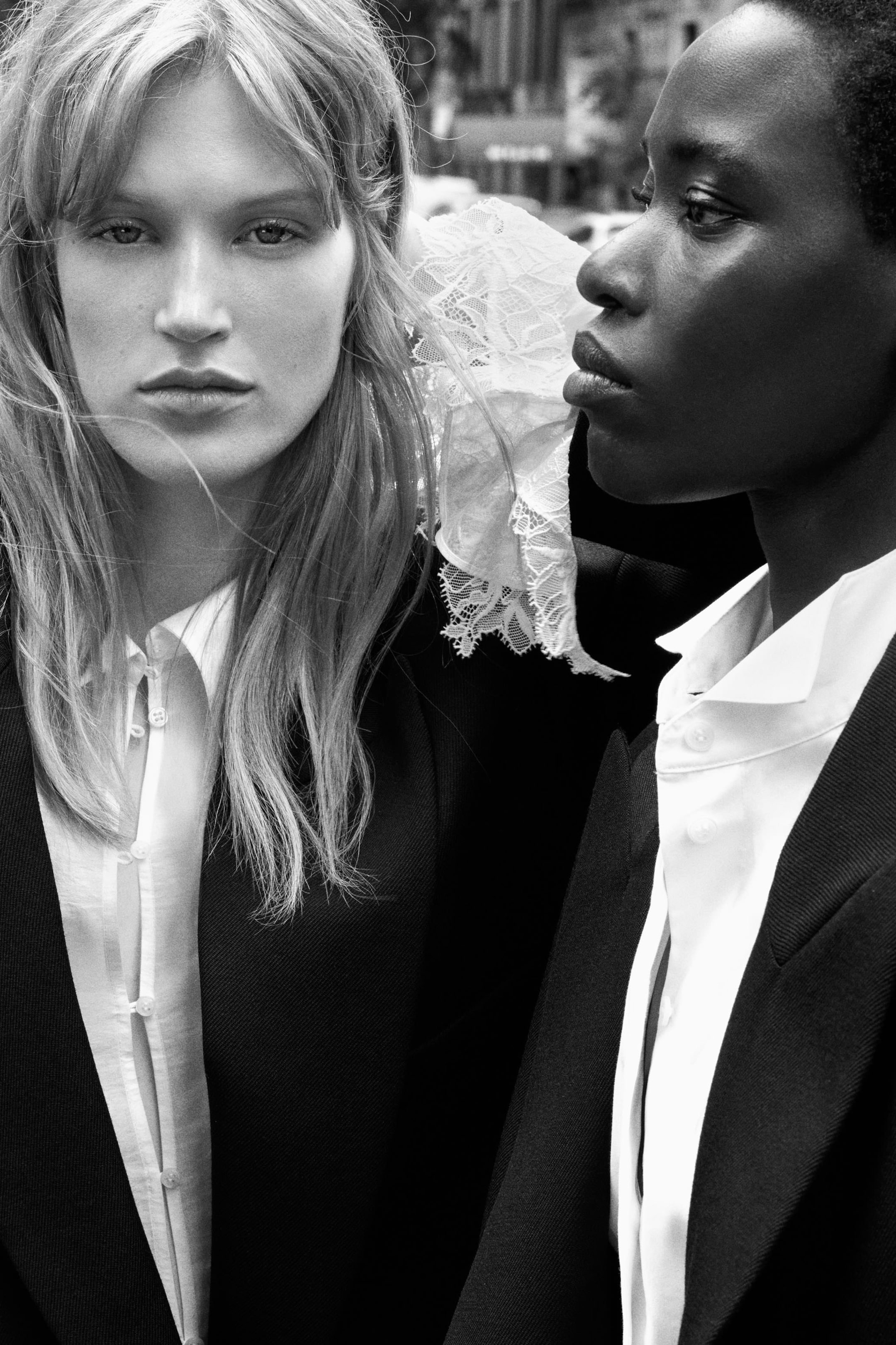 Two people in black and white, wearing suits and white shirts. One has long hair; the other has short hair. The setting appears urban.