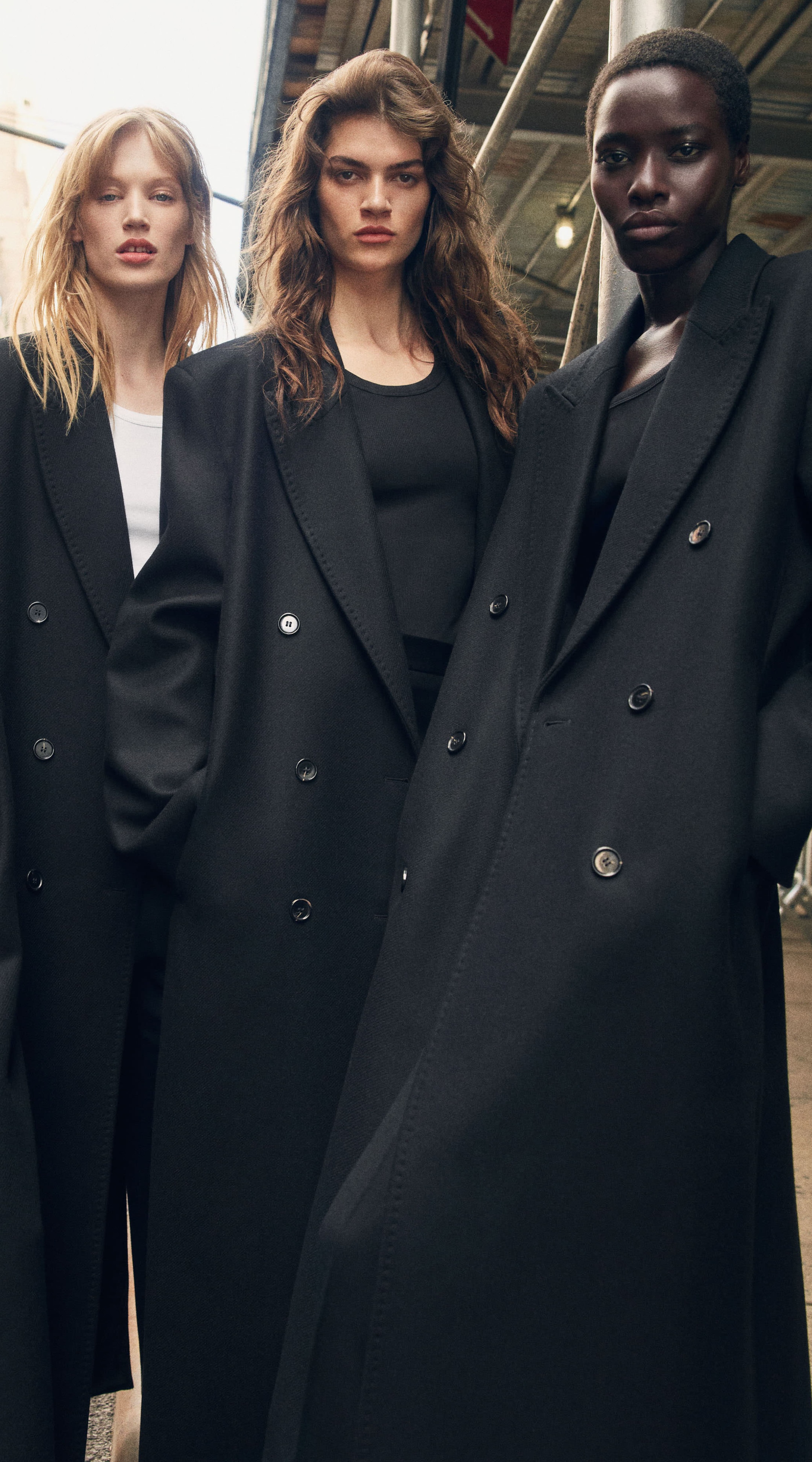 Three models in stylish long black coats stand confidently on a city street, each with a distinct expression.