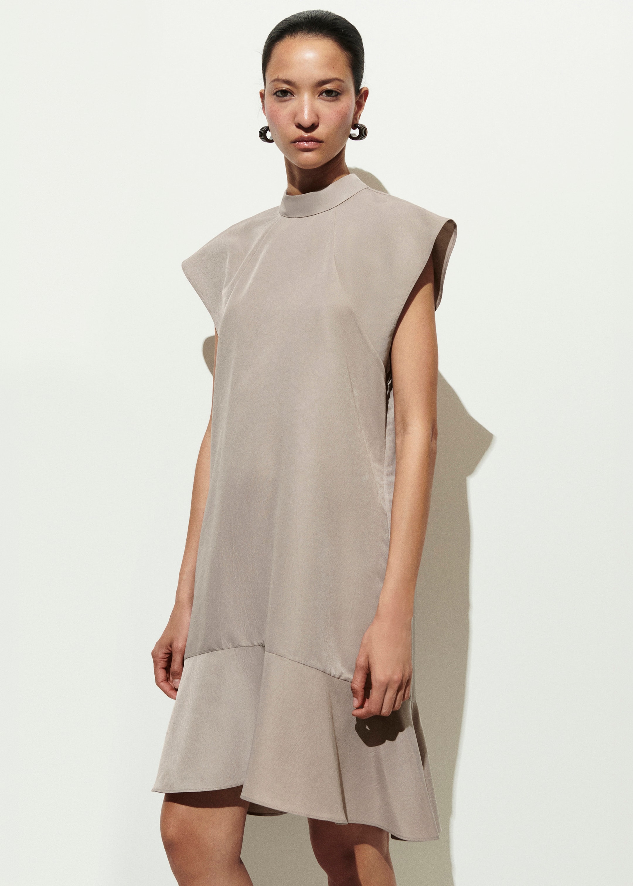 Woman in a taupe sleeveless dress with shoulder pads, standing against a white background, wearing hoop earrings.