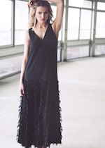 Placeholder for: Woman in a black sleeveless dress with feather details stands in a bright, spacious room with large windows, hand resting on her head.