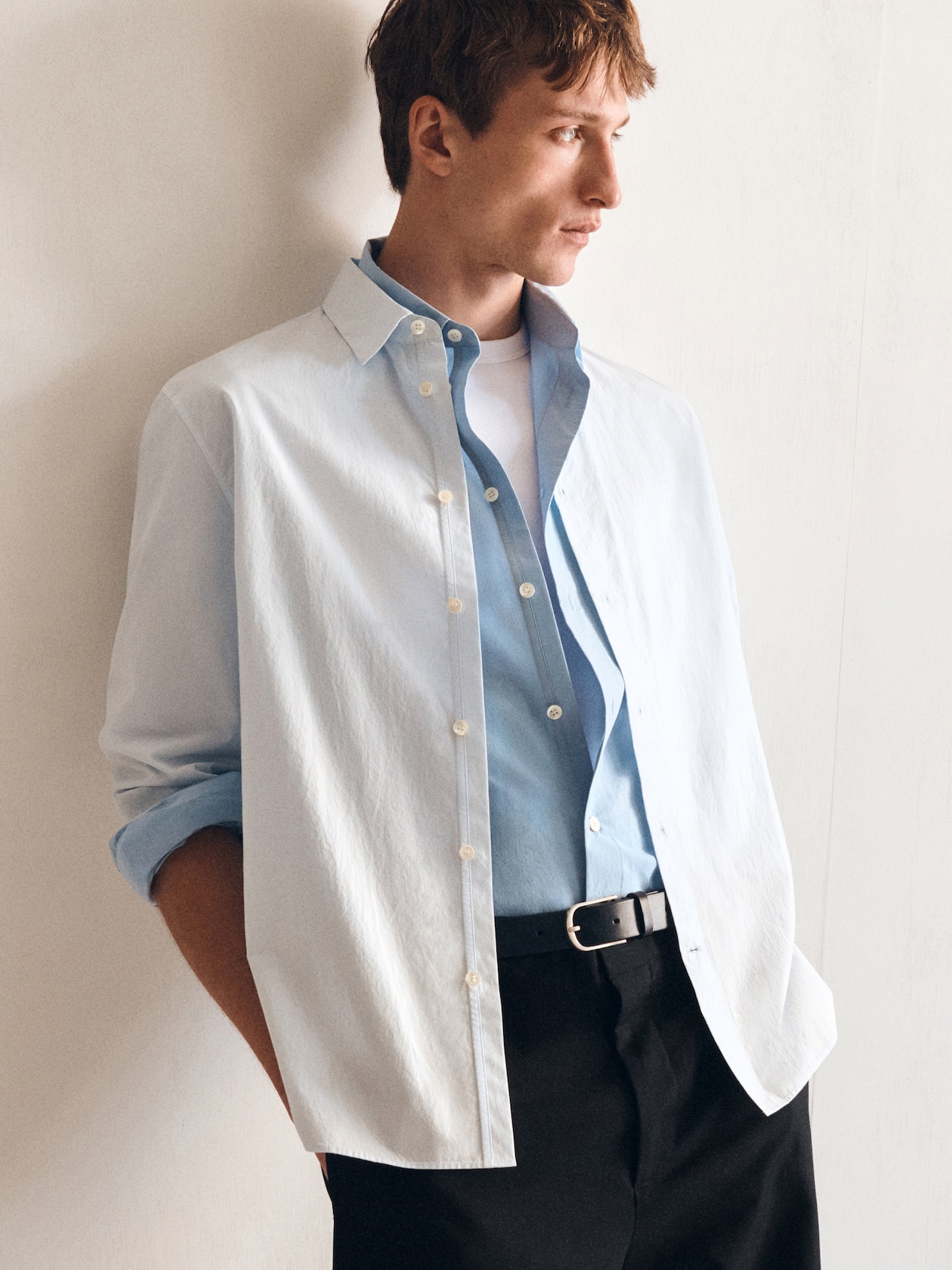Man in a casual outfit with a white and blue shirt layered over a white tee, standing against a light-colored wall, looking to the side.