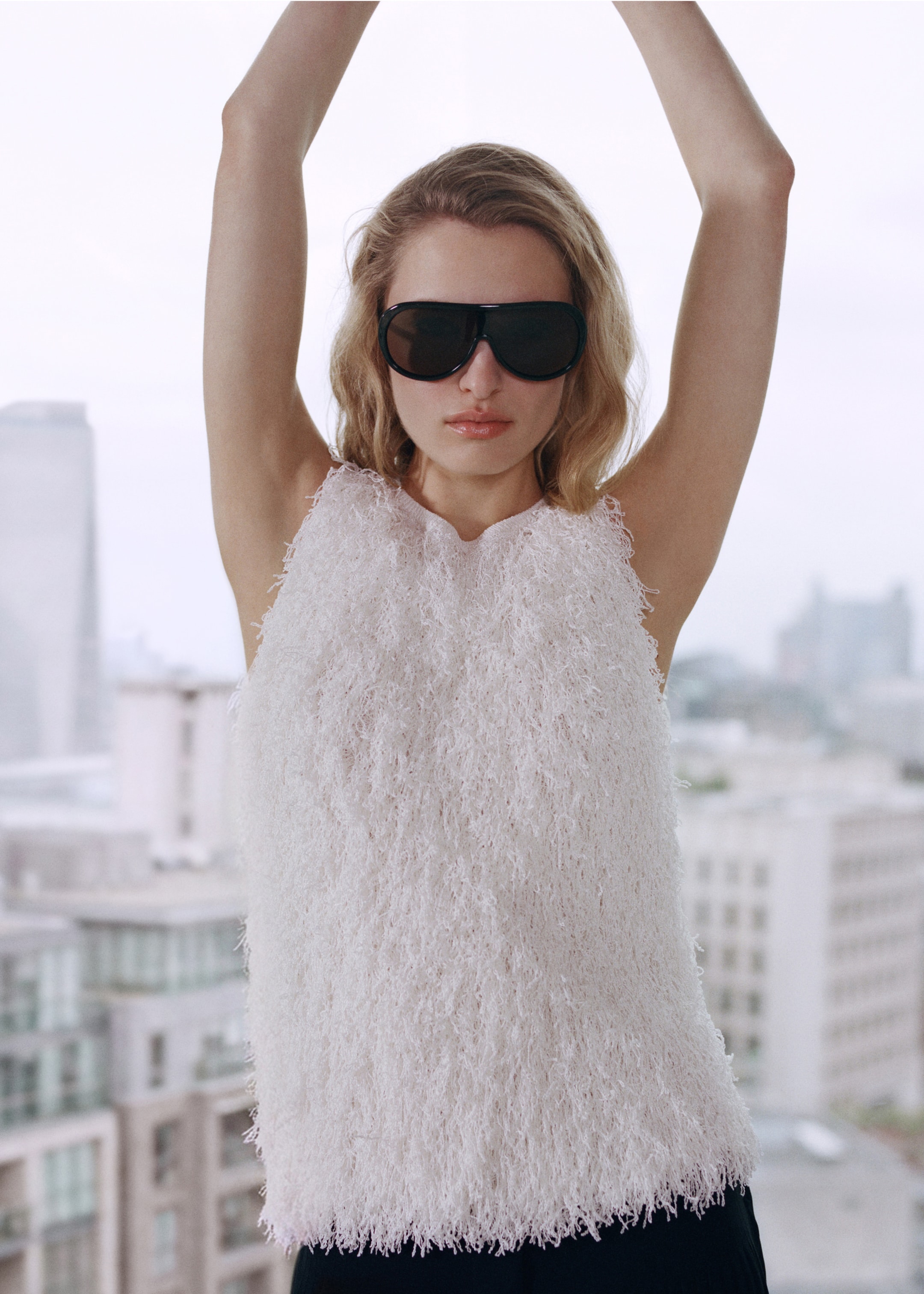 A person with long hair and sunglasses, wearing a textured white top, stands with arms raised against a cityscape backdrop.