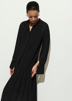 Placeholder for: Person in a sleek black dress with pleated details, looking down, standing against a light background.