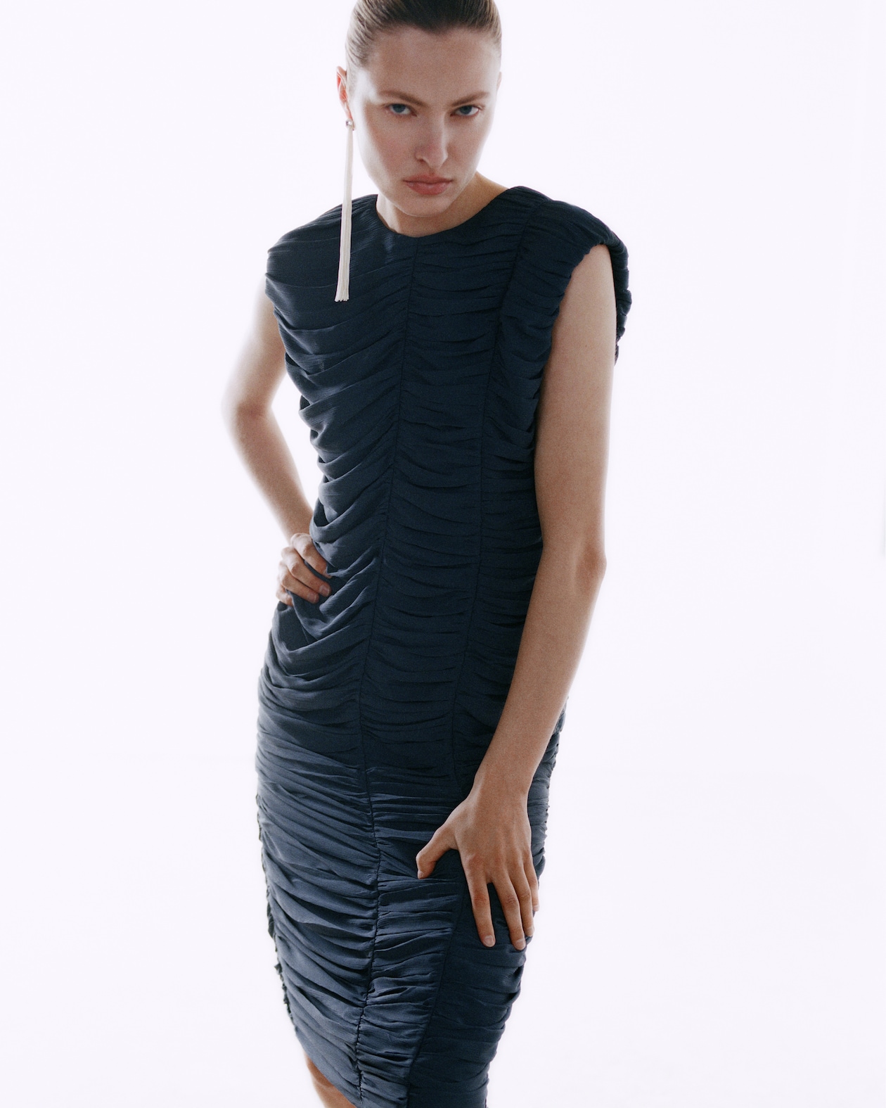 Person in a fitted, ruched black dress with a long, single earring, standing confidently against a plain white background.