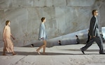 Placeholder for: Models in flowing outfits and suits walk in a line on a textured concrete surface, against a large, angular stone backdrop.