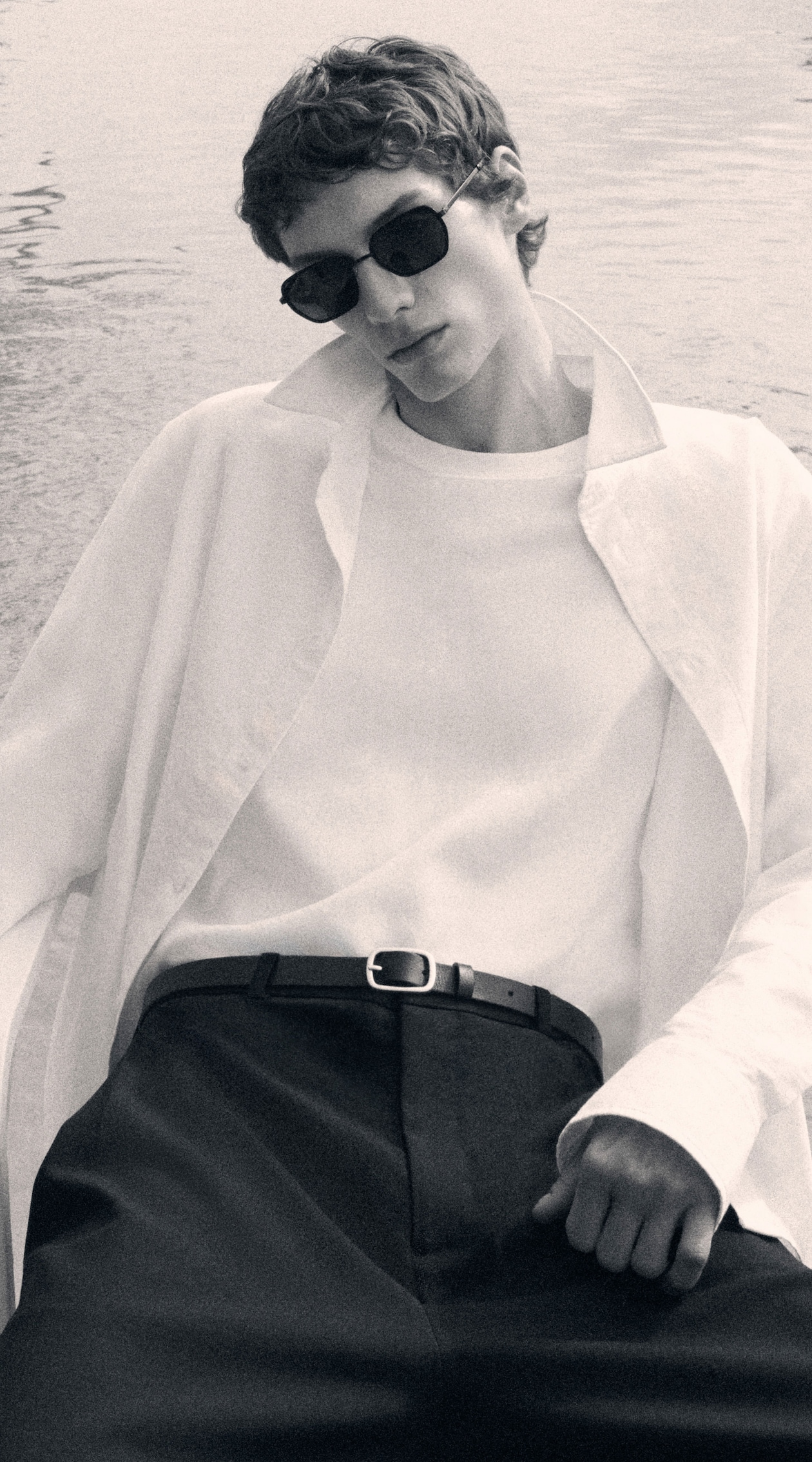 Person in a relaxed pose wearing sunglasses, a white shirt, white undershirt, and dark pants. Background appears serene and watery.