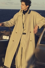 Placeholder for: Person in a beige trench coat and suit leans against a vintage car by the ocean on a sunny day.