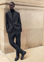 Placeholder for: A person in a black suit and turtleneck leans casually against a beige stone wall, with hands in pockets and legs crossed on a sidewalk.