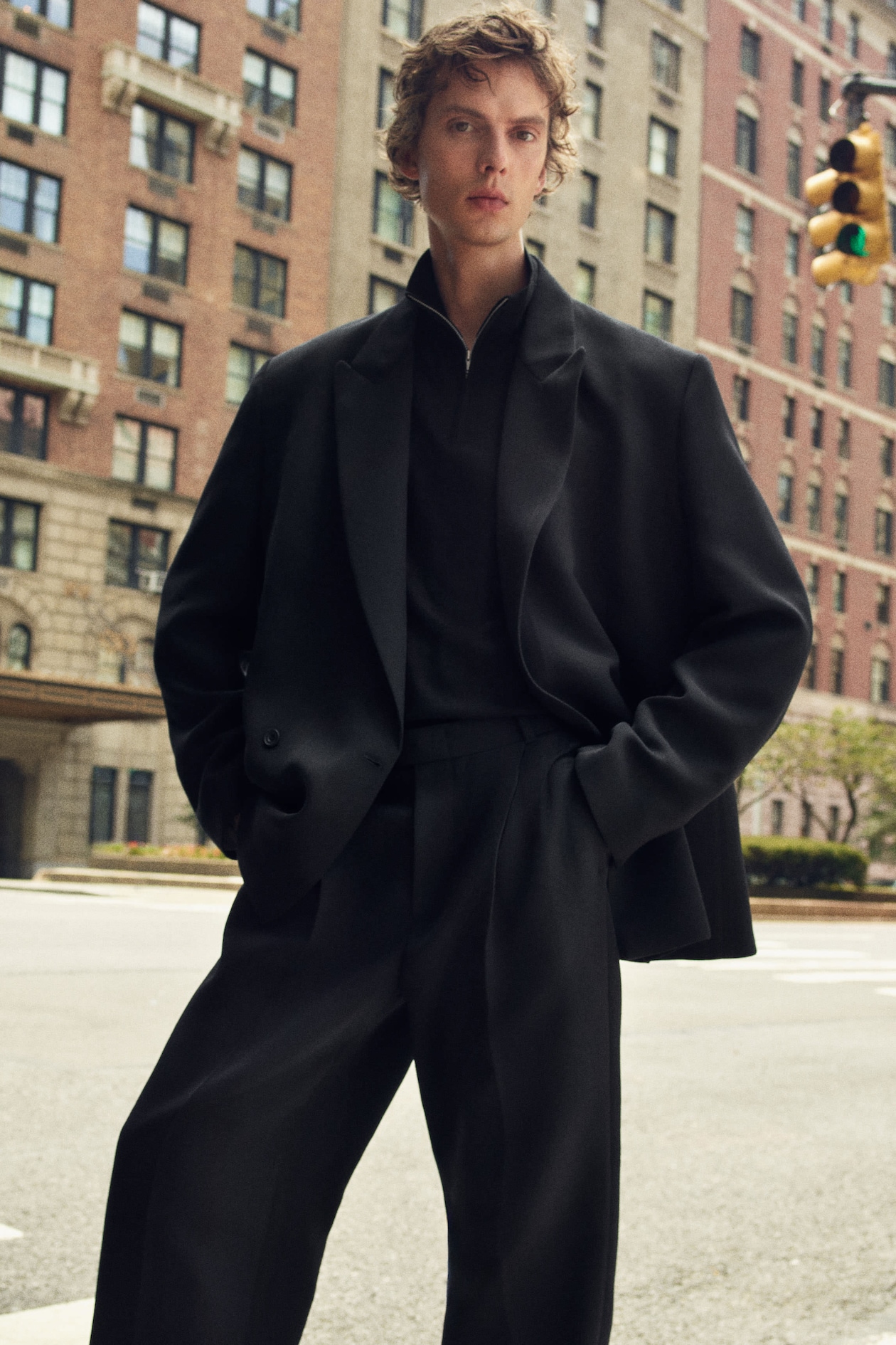 A person with curly hair stands confidently on a city street, wearing an oversized black suit, with tall buildings in the background.