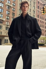 Placeholder for: A person with curly hair stands confidently on a city street, wearing an oversized black suit, with tall buildings in the background.