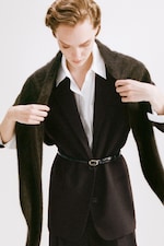 Placeholder for: Person in a brown suit and white shirt adjusts a dark shawl draped over their shoulders, with a belt cinching the waist.