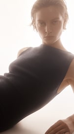 Placeholder for: Person with short hair in a sleeveless, textured dress, leaning slightly back against a soft light background, gazing directly ahead.