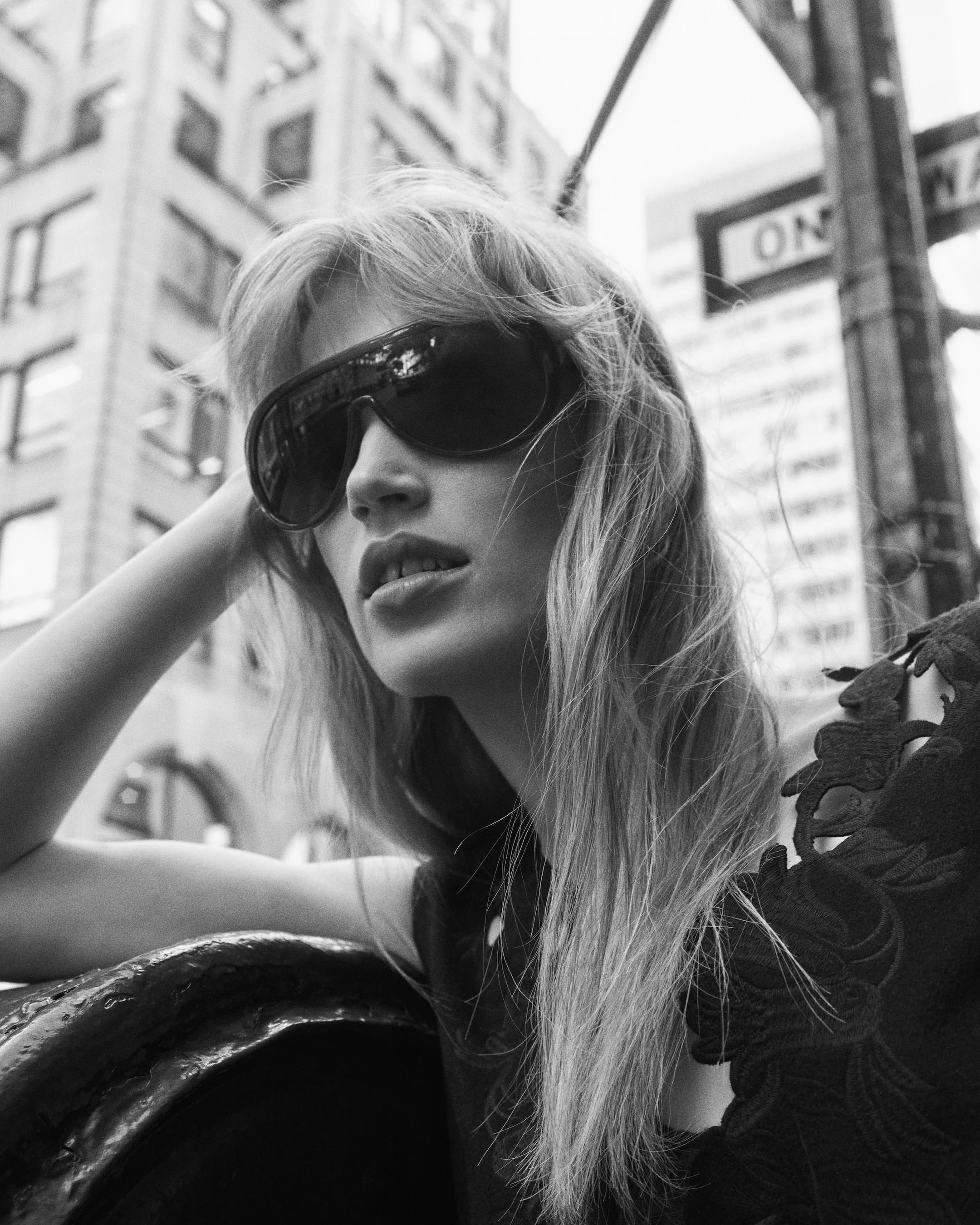 Black and white photo of a woman with long hair wearing large sunglasses, leaning on a railing, with city buildings in the background.