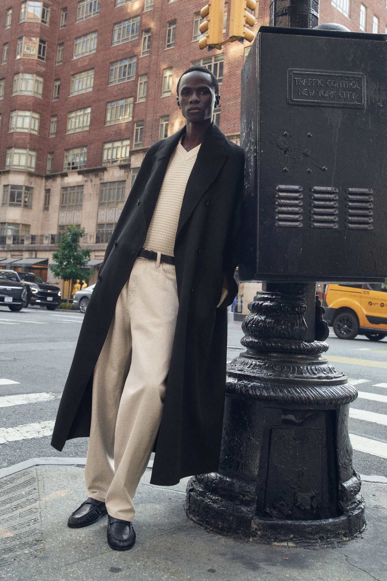 Person in a long dark coat and beige outfit leans against a traffic control box on a New York City street, with yellow taxis and buildings in the background.