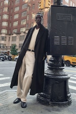 Placeholder for: A person leans against a traffic control box on a New York City street corner, wearing a long coat and neutral-toned clothing against a backdrop of buildings.