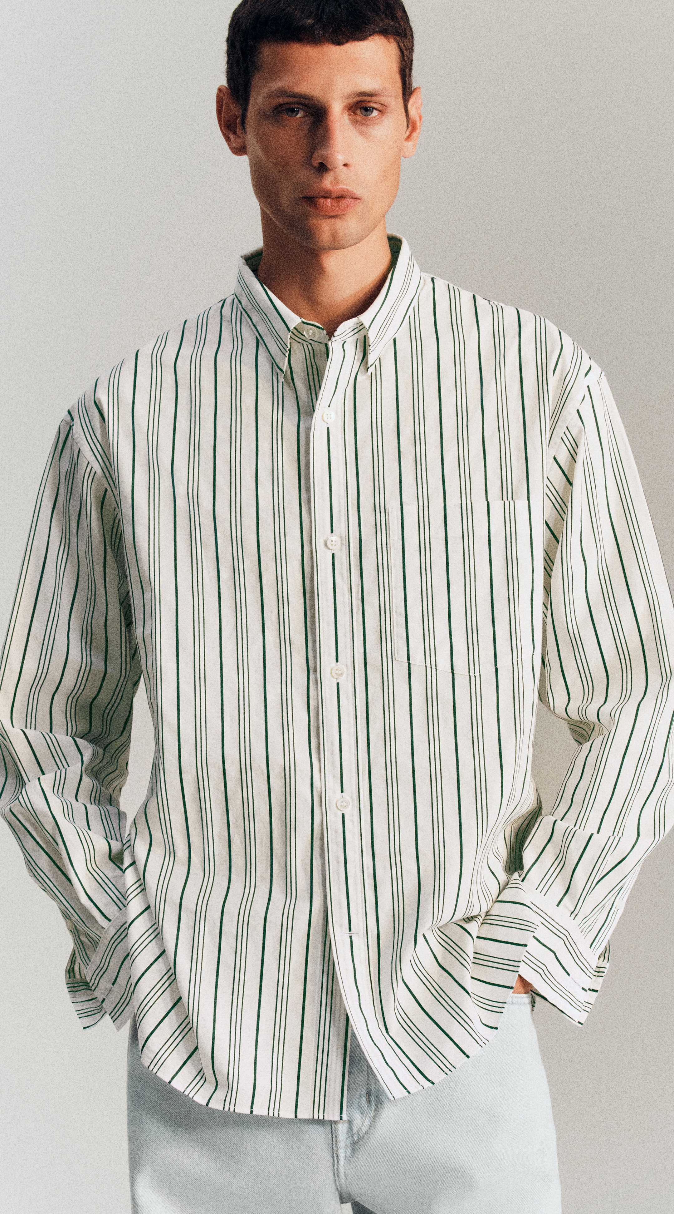 Man wearing a white shirt with green stripes, standing with hands in light pants pockets.