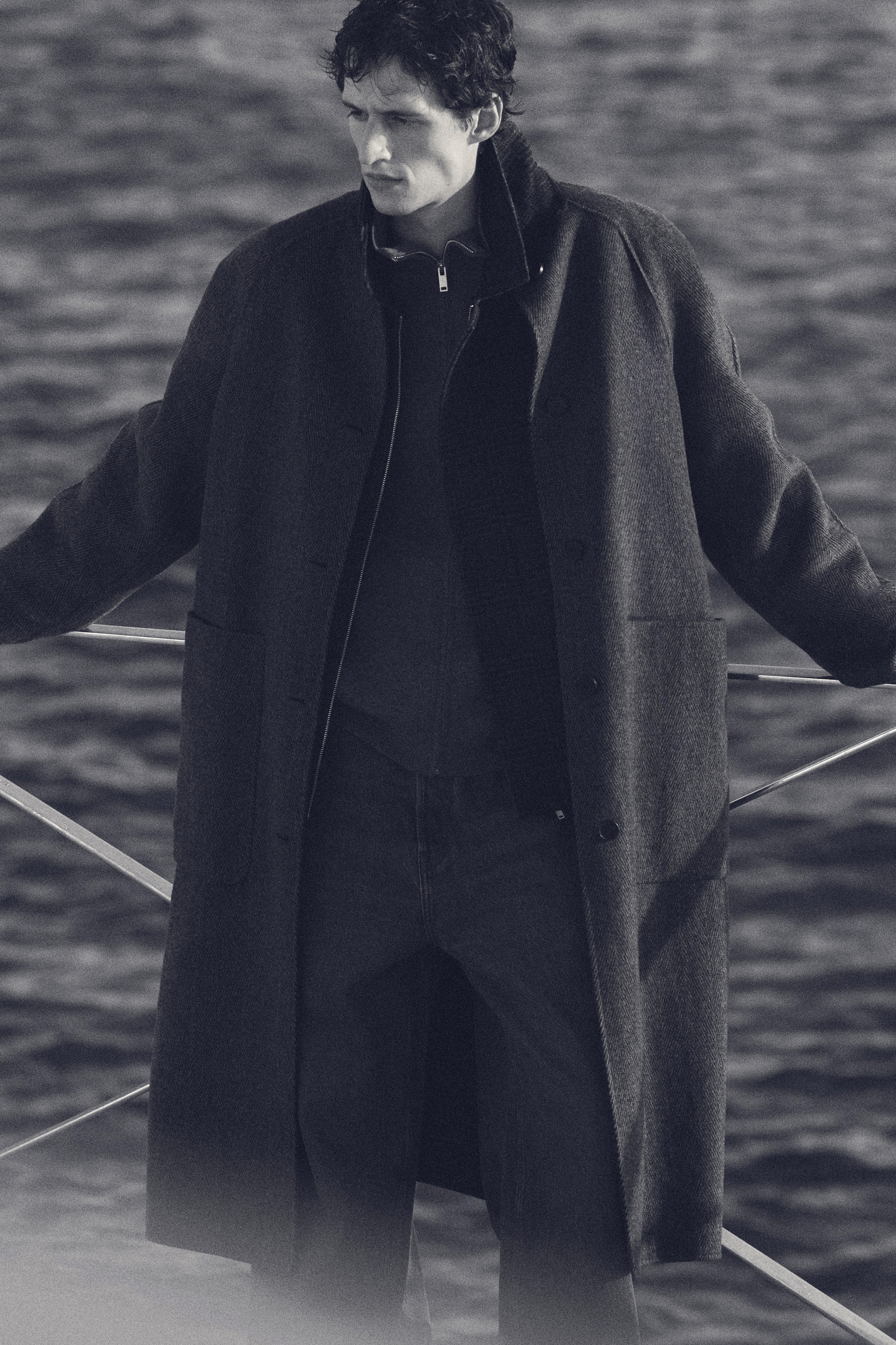 A person in a long coat stands by a railing, gazing over a body of water. The scene is in black and white.