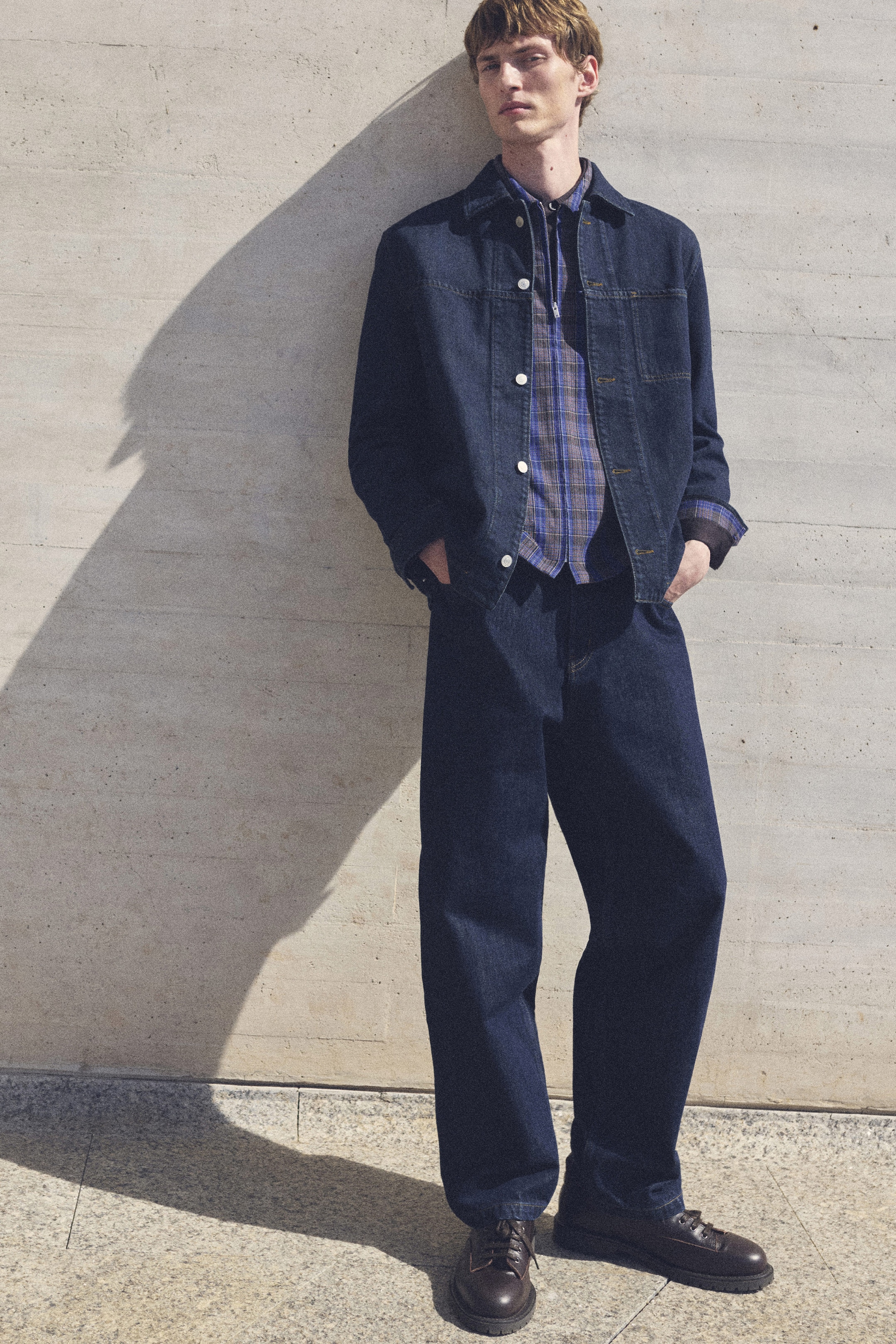 A person in a denim jacket and jeans stands against a light stone wall, hands in pockets, casting a shadow.