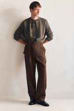 Placeholder for: Man in a gray sweater and brown high-waisted trousers leans against a white wall, looking to the side, hands behind his back.