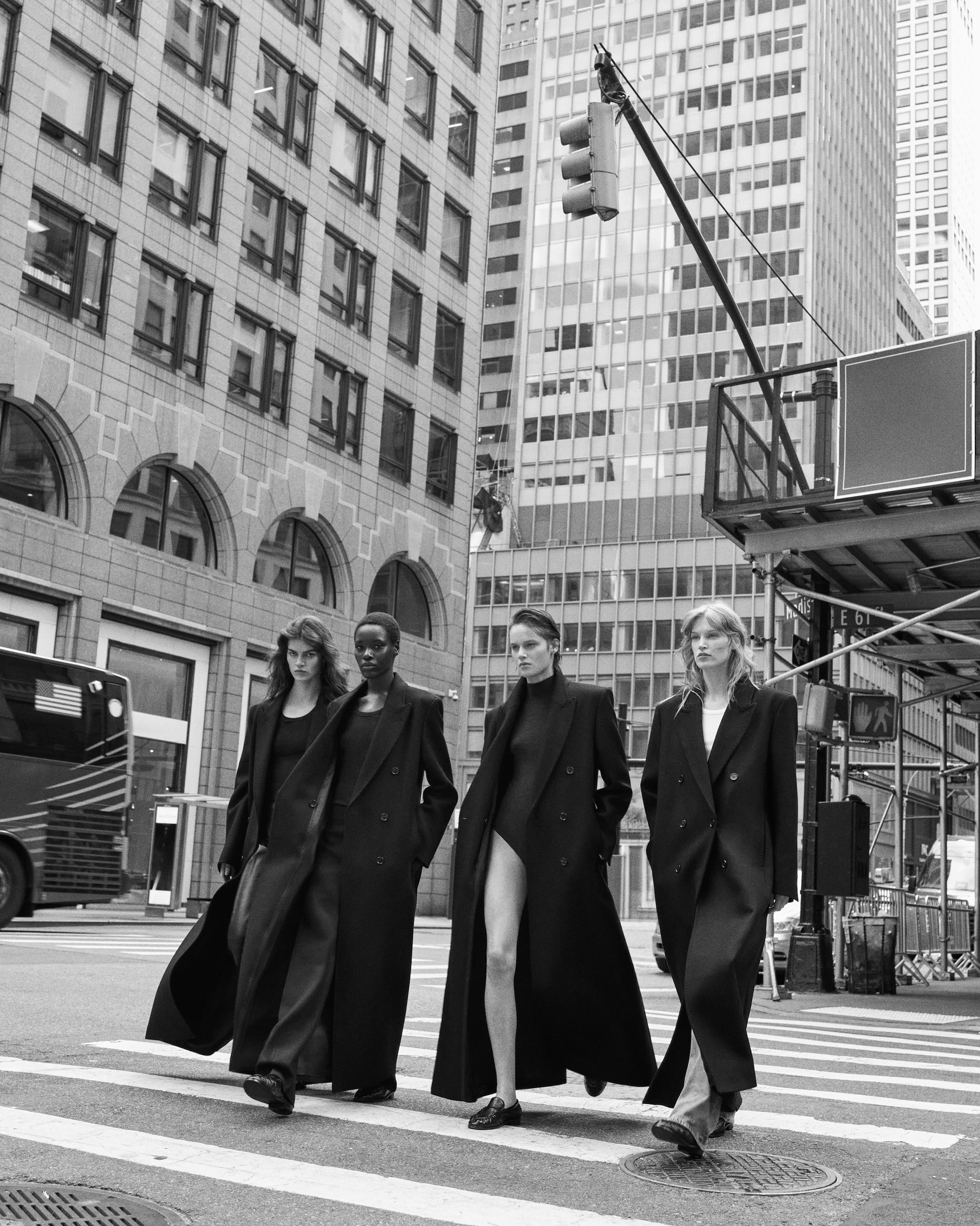 Four people in long coats walk confidently across a city street. Urban buildings and a traffic light frame the scene in black and white.