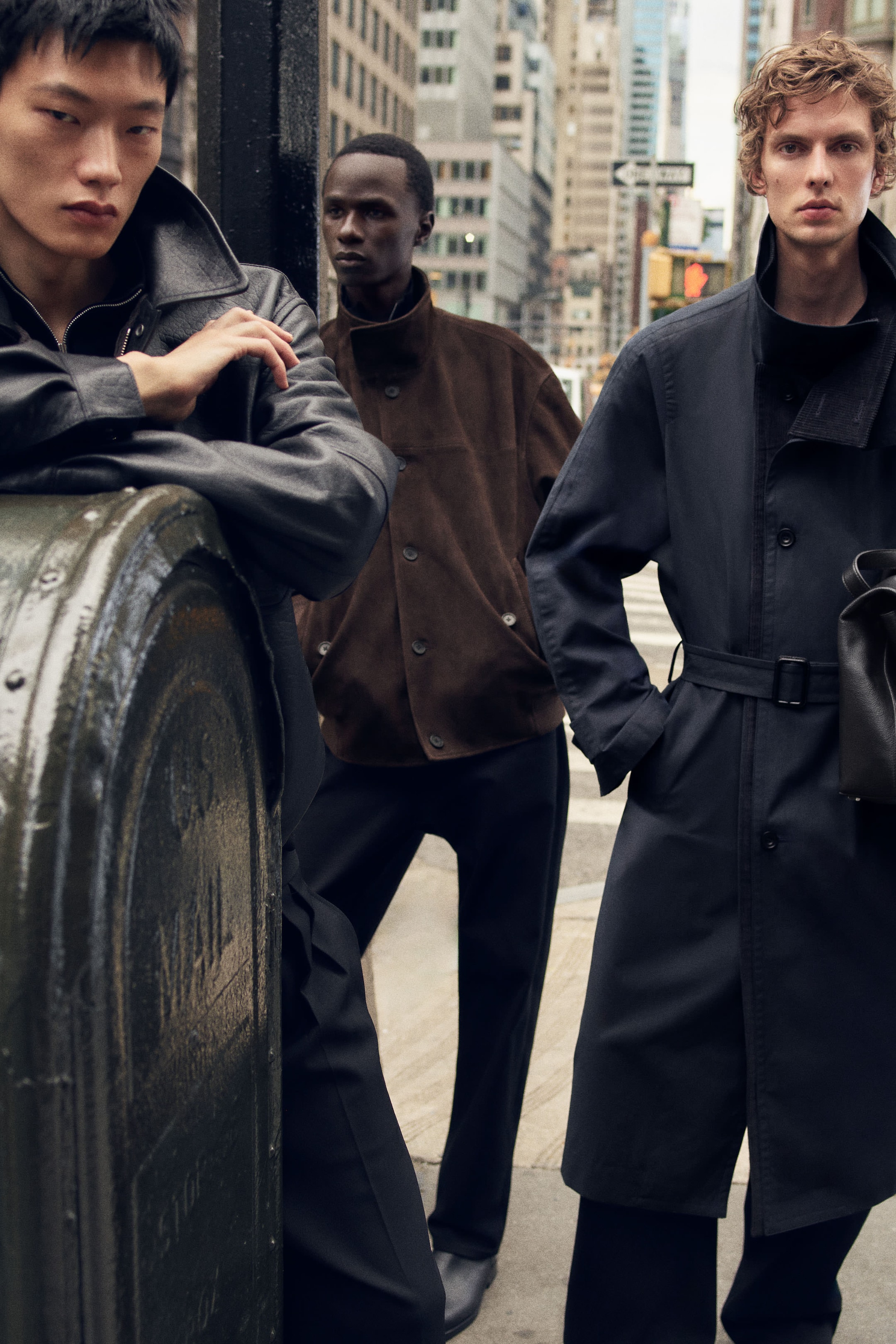 Three men in stylish coats stand on a city street, with tall buildings in the background, creating a fashionable urban scene.