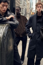 Placeholder for: Three men in stylish coats stand on a city street, with tall buildings in the background, creating a fashionable urban scene.