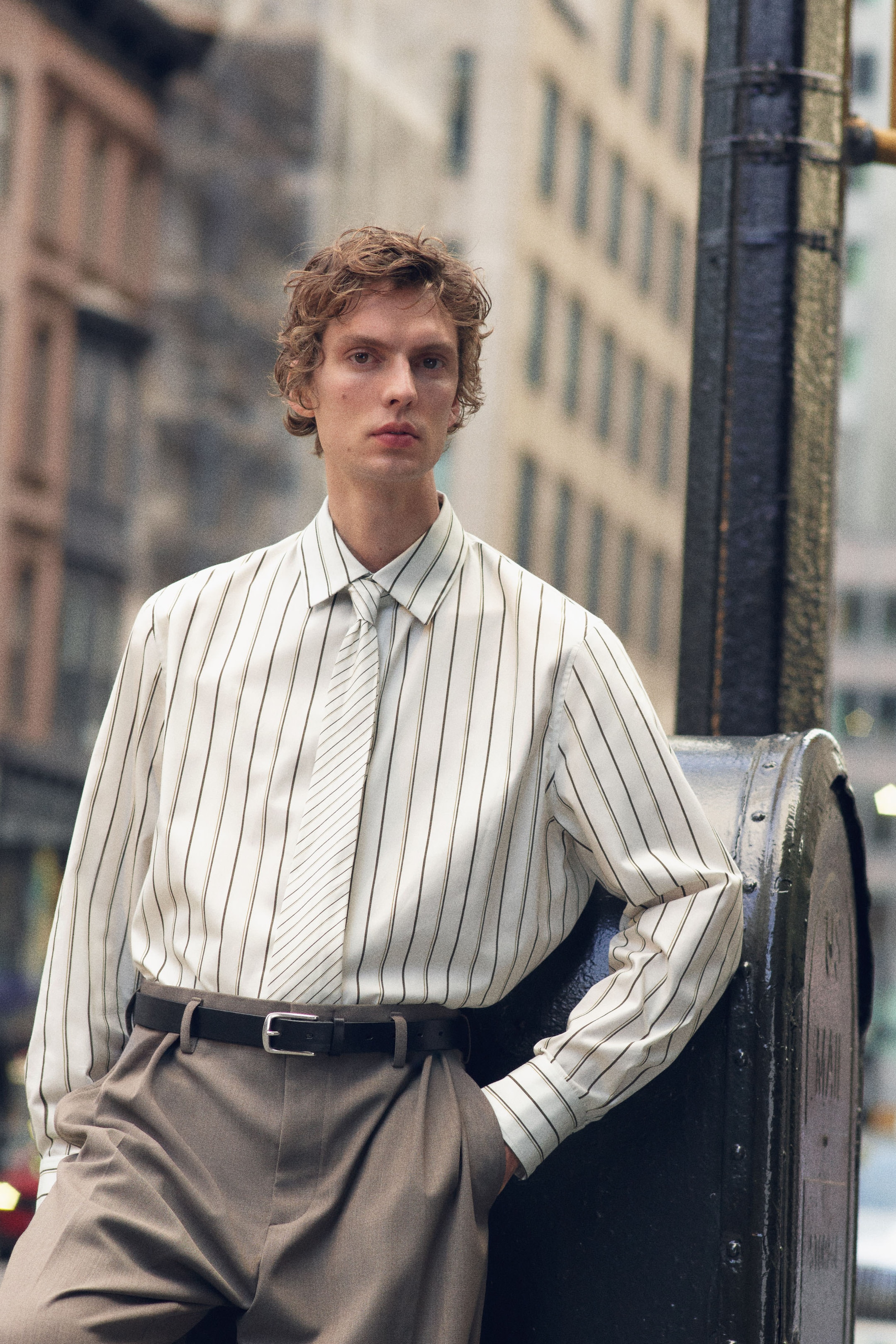 A person in a striped white shirt and beige trousers leans against a metal post on a city street, with blurred buildings in the background.