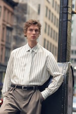 Placeholder for: A person in a striped white shirt and beige trousers leans against a metal post on a city street, with blurred buildings in the background.
