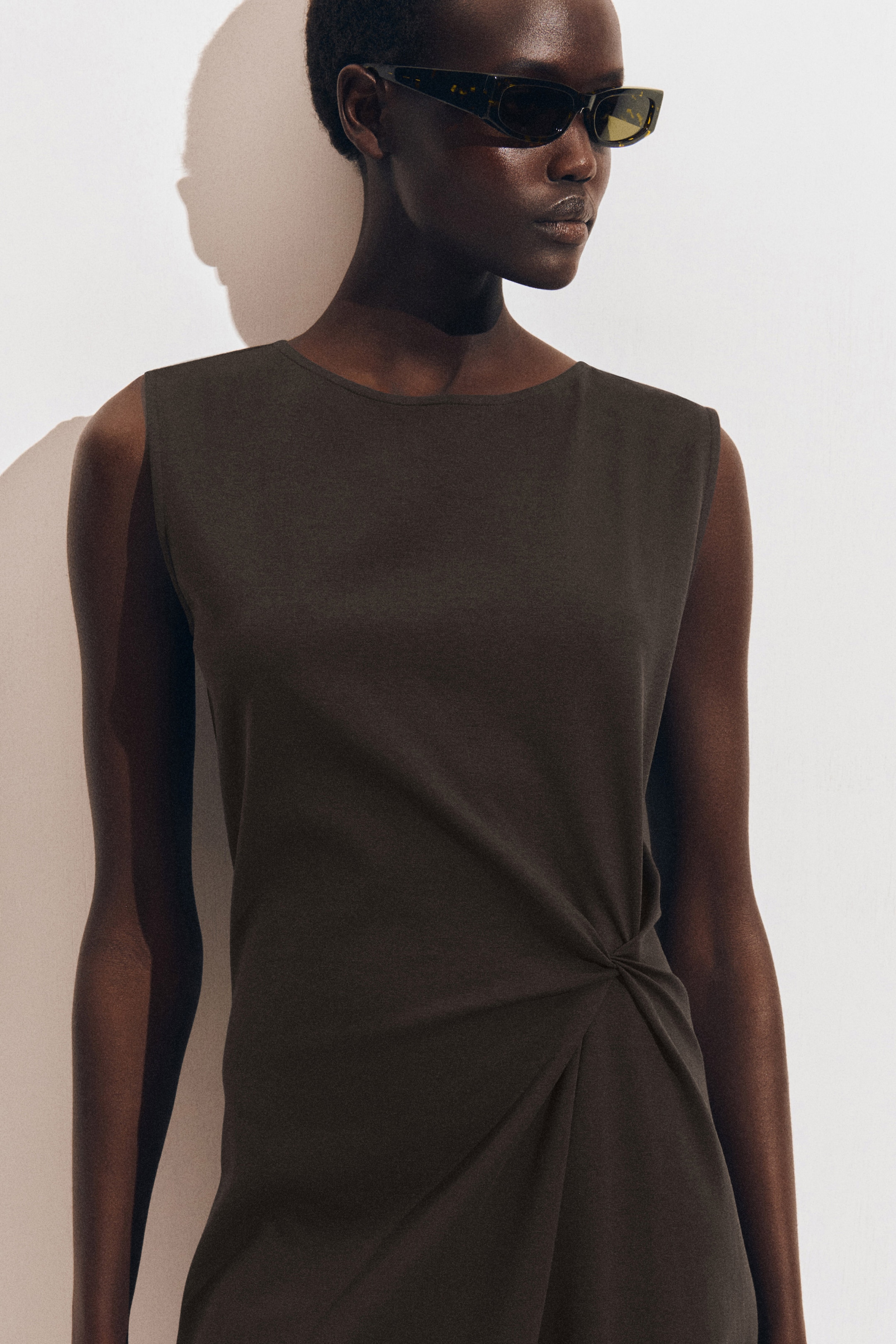 Person wearing dark sunglasses and a sleeveless black dress with a gathered design, standing against a plain white background.