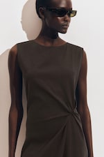 Placeholder for: Person wearing dark sunglasses and a sleeveless black dress with a gathered design, standing against a plain white background.