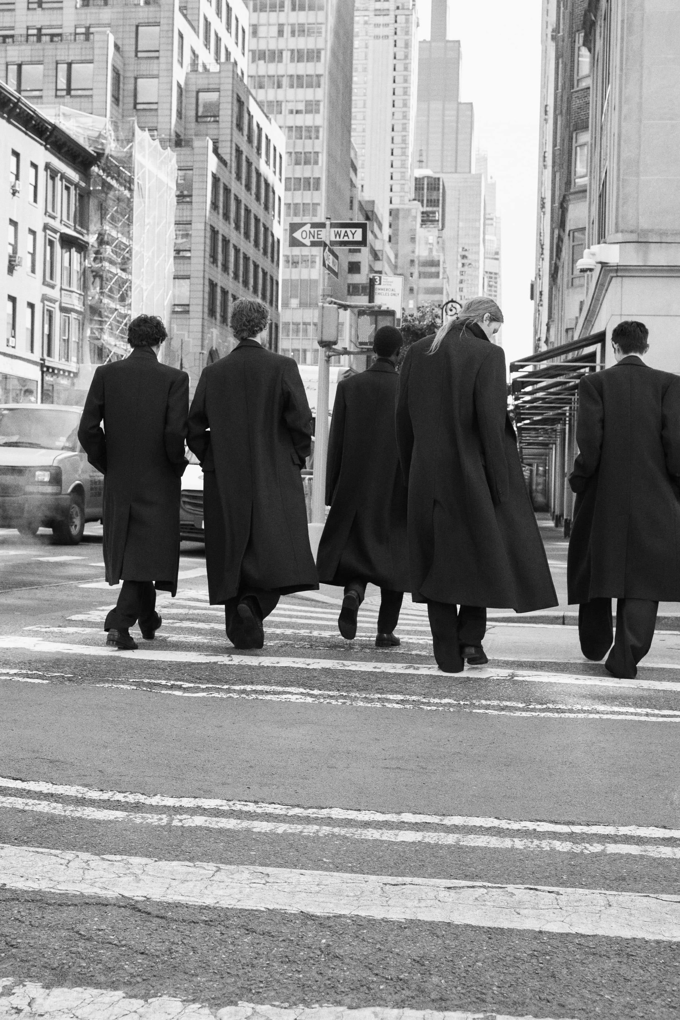 Five people in long coats walking in a city street crosswalk, surrounded by tall buildings on a bright day.