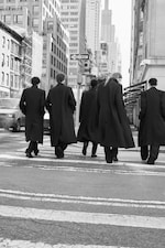 Placeholder for: Five people in long coats walking in a city street crosswalk, surrounded by tall buildings on a bright day.