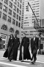Placeholder for: Four women in long black coats walk confidently across a city street, surrounded by tall buildings.