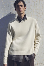 Placeholder for: Man in a white sweater over a dark shirt stands against a textured white wall, casting a shadow.