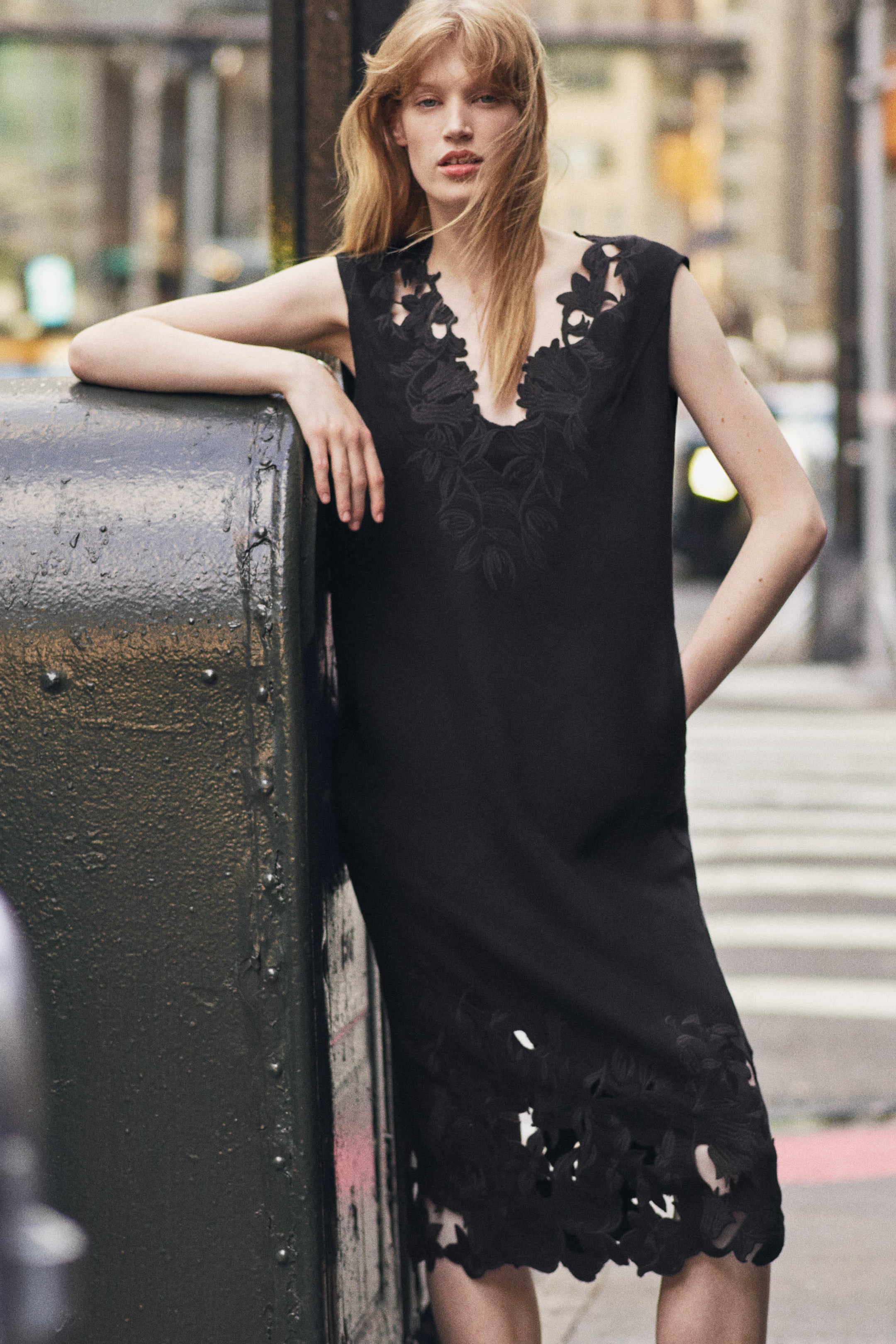 A woman with long hair poses in a sleeveless black dress with floral cutouts, leaning against a metal surface in an urban setting.