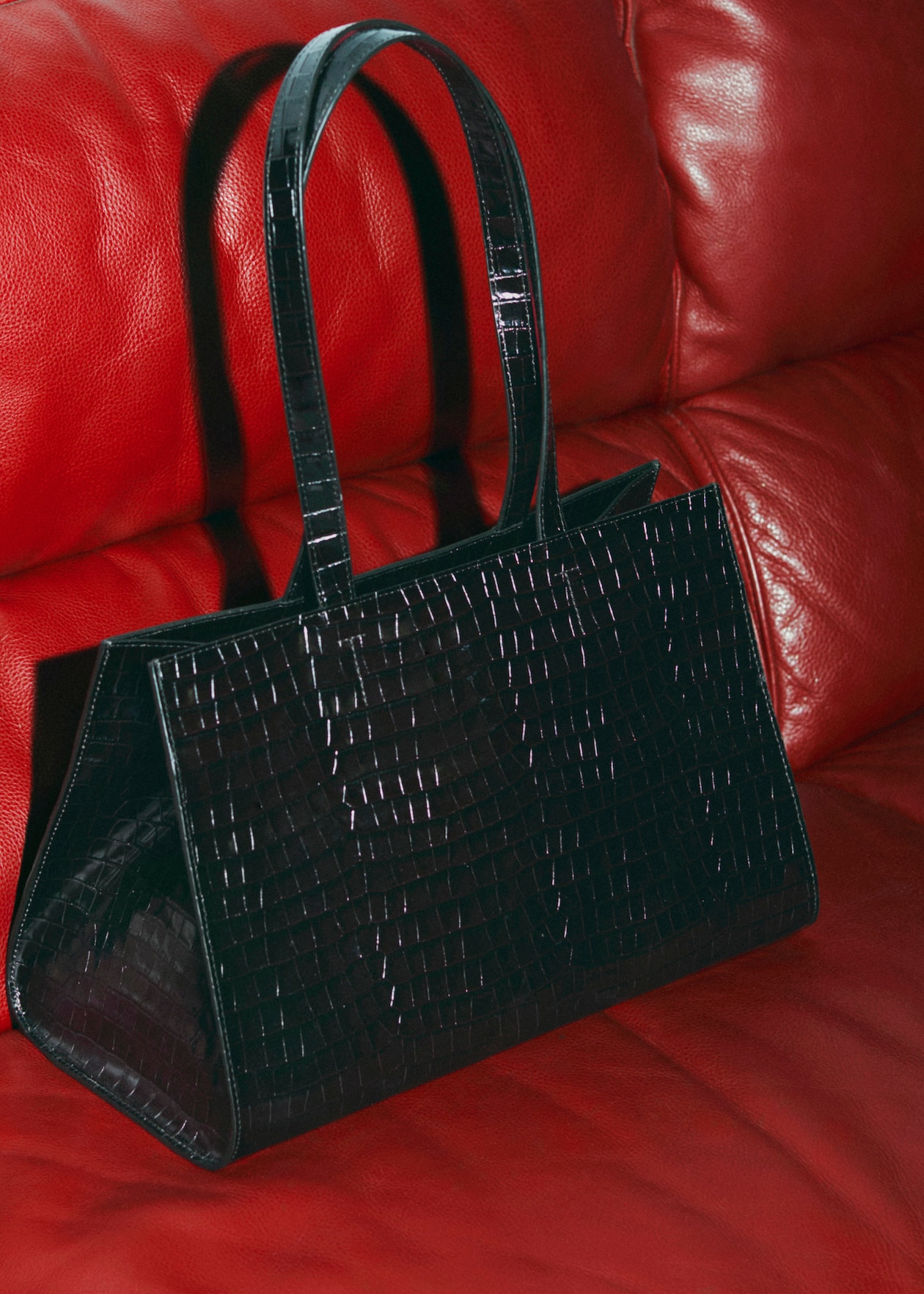 Black crocodile-patterned handbag with long handles on a red leather couch.