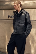 Placeholder for: Person in a black leather jacket and black pants stands against a stone wall with the word "Paris" partially visible.