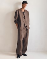 Placeholder for: A person stands against a plain wall wearing an oversized brown suit and black shoes, with hands in pockets.