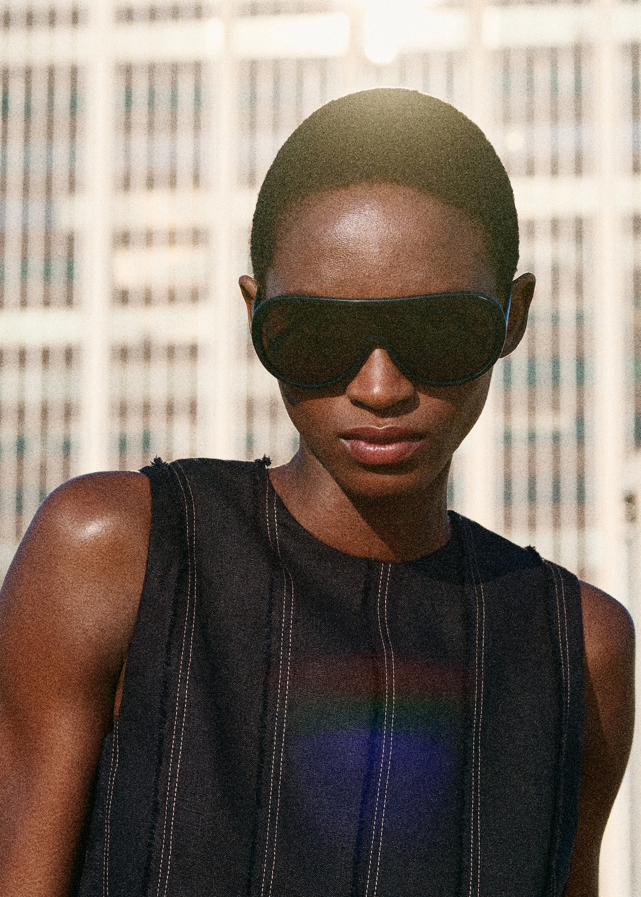 A person wearing large sunglasses and a sleeveless top stands outdoors with a building in the background under bright sunlight.