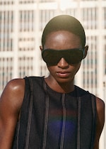 Placeholder for: A person wearing large sunglasses and a sleeveless top stands outdoors with a building in the background under bright sunlight.