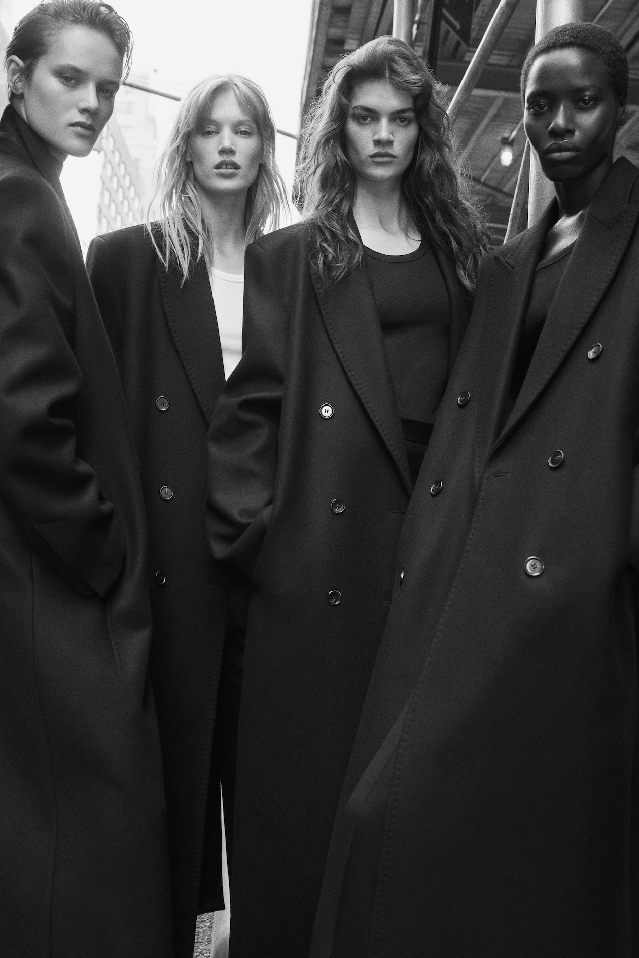 Four models wearing long, dark overcoats stand confidently on an urban street, captured in a monochromatic style.