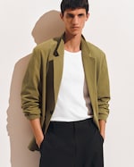 Placeholder for: Person wearing an olive blazer, white shirt, and black pants, standing against a light background with hands in pockets.