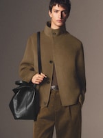 Placeholder for: A person wearing an olive green coat, brown pants, and holding a black leather bag, stands against a neutral background.