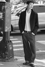 Placeholder for: A person in a fashionable outfit stands on a city street, leaning against a post, with hands in pockets and a car in the background.