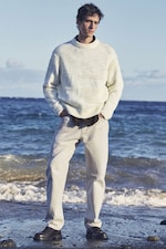 Placeholder for: Man wearing a white sweater and light jeans stands on a beach, hands in pockets, with the ocean in the background under a clear sky.