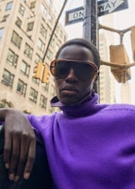 Placeholder for: A person wearing large sunglasses and a purple turtleneck stands in front of city buildings and a one-way sign.
